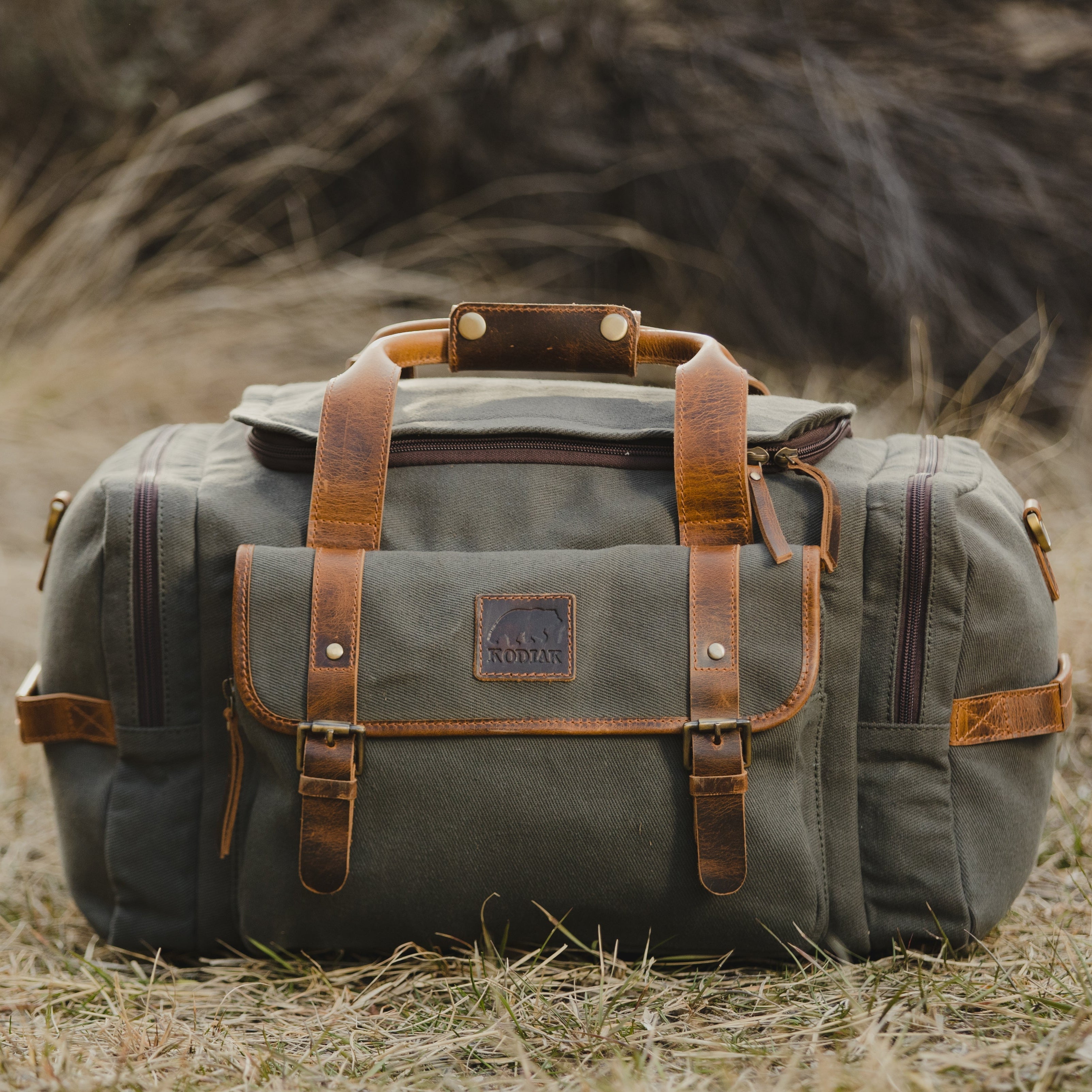 Green canvas duffel bag with brown leather straps on a grassy field