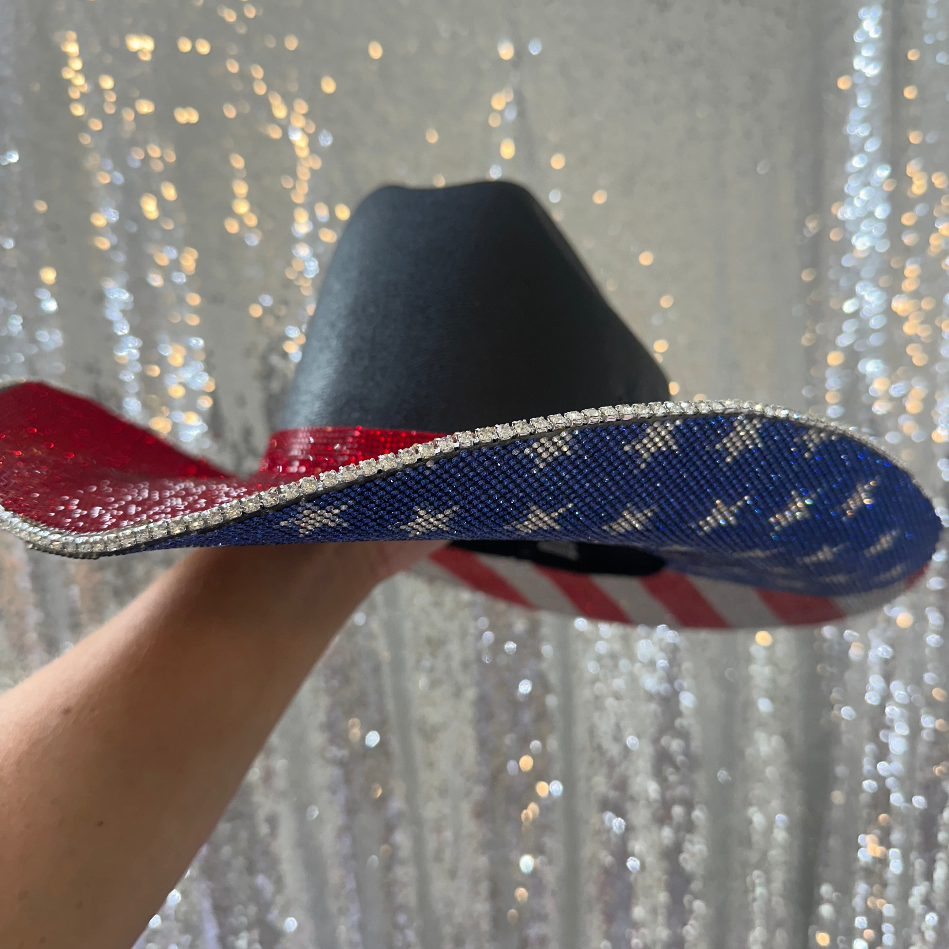 Hand holding a decorative hat with red, white, and blue colors against a glittery silver background.
