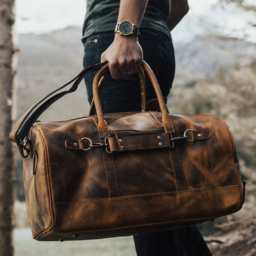 Person holding a brown leather bag in a natural setting with trees and water.