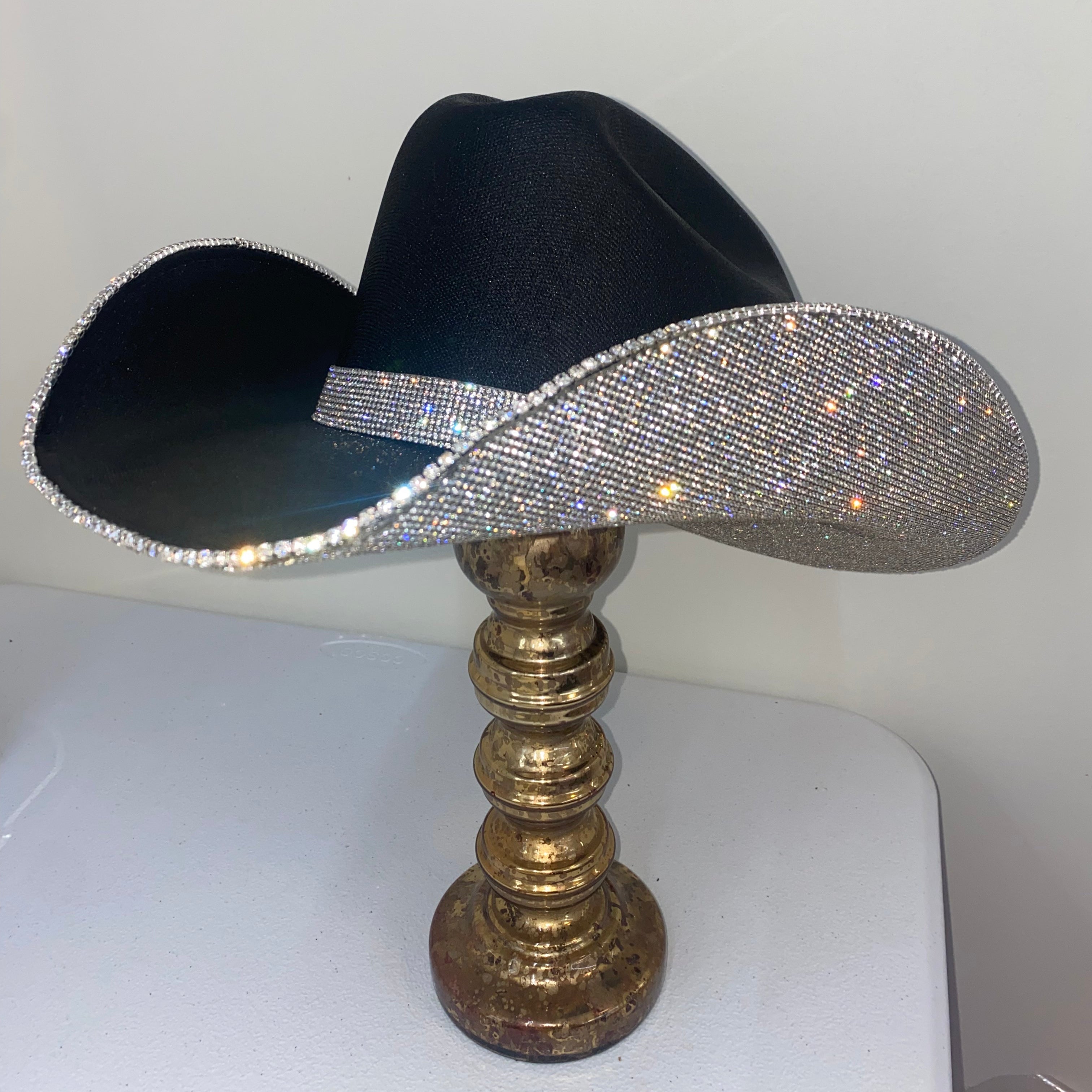 Black cowboy hat with a rhinestone band on a gold stand against a white background