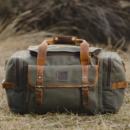 Green canvas duffel bag with brown leather straps on a grassy field