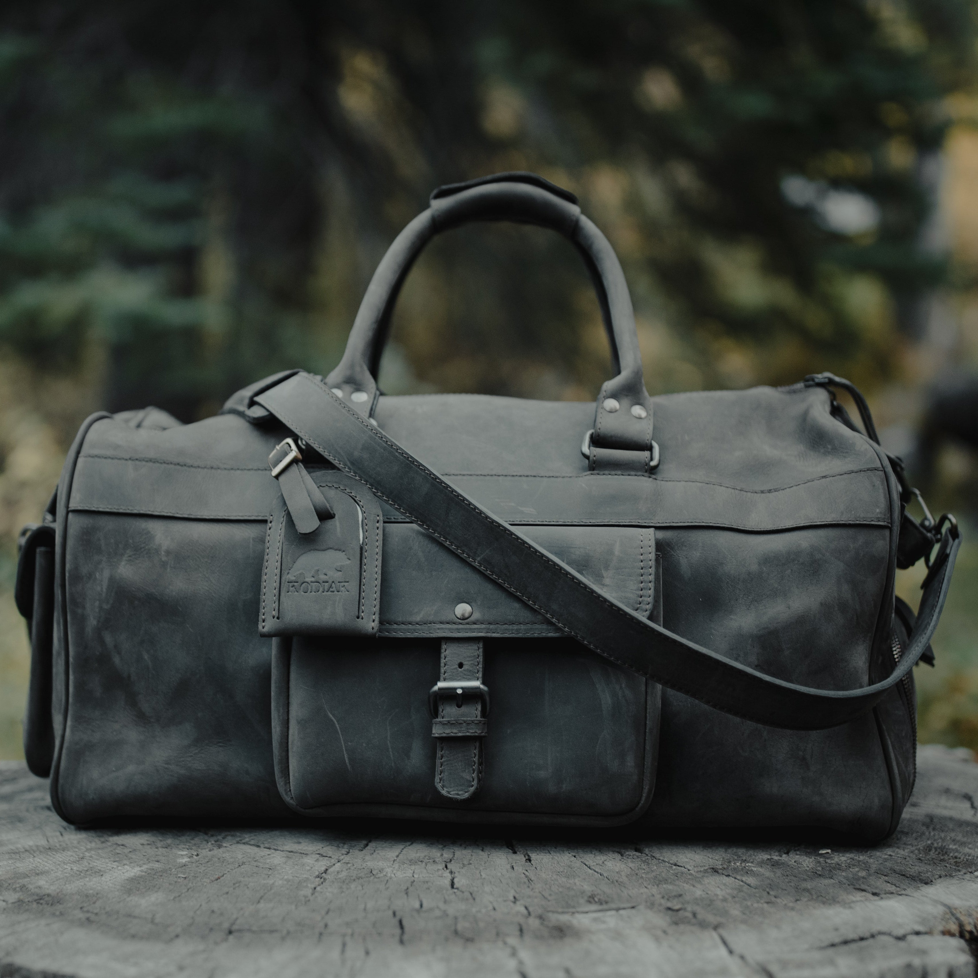 Dark gray leather duffel bag on a wooden stump with a blurred natural background