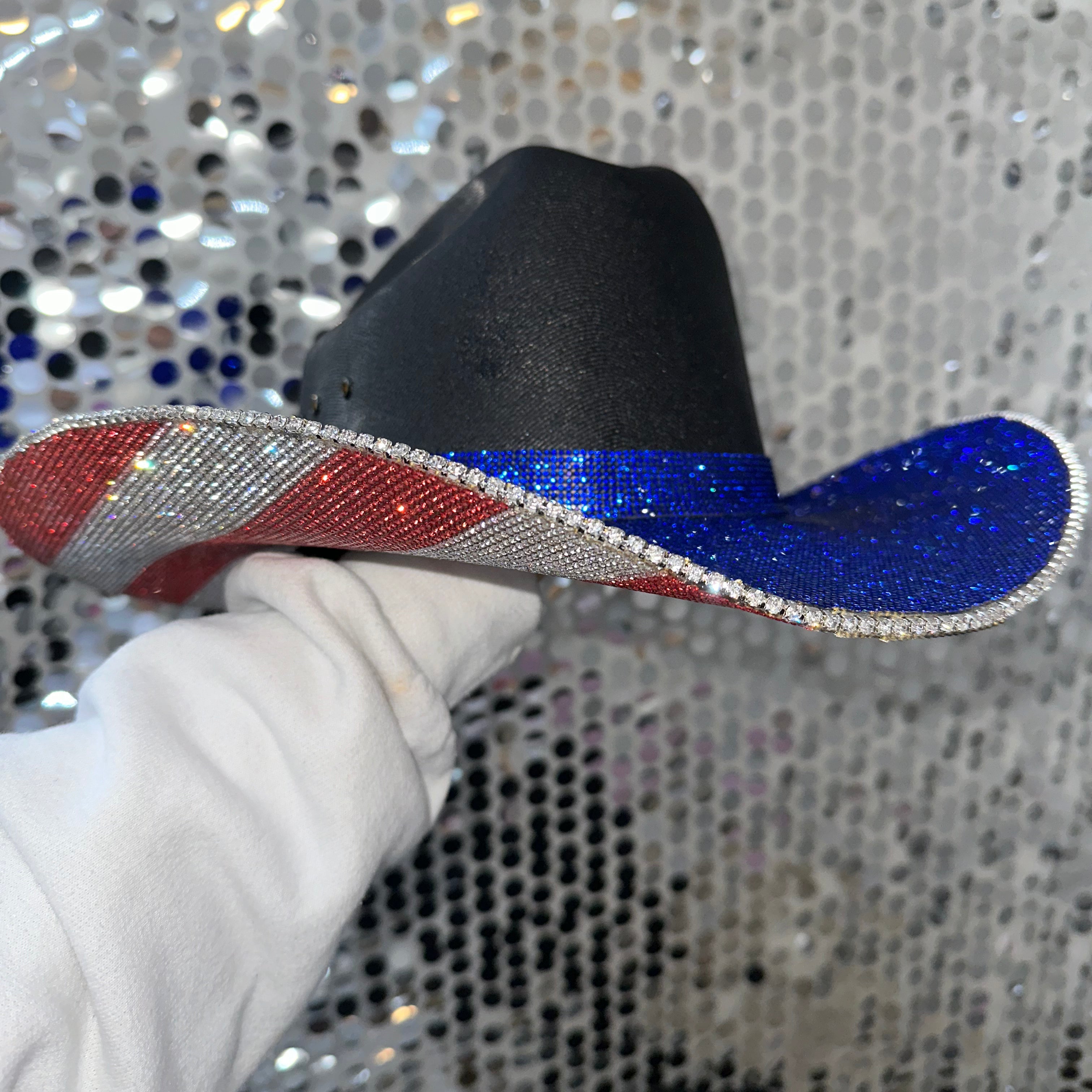 Black cowboy hat with red and blue glittery brim held by a gloved hand against a silver sequin background.
