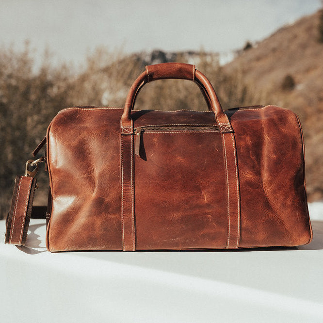 Brown leather duffel bag on a white surface with a natural landscape background