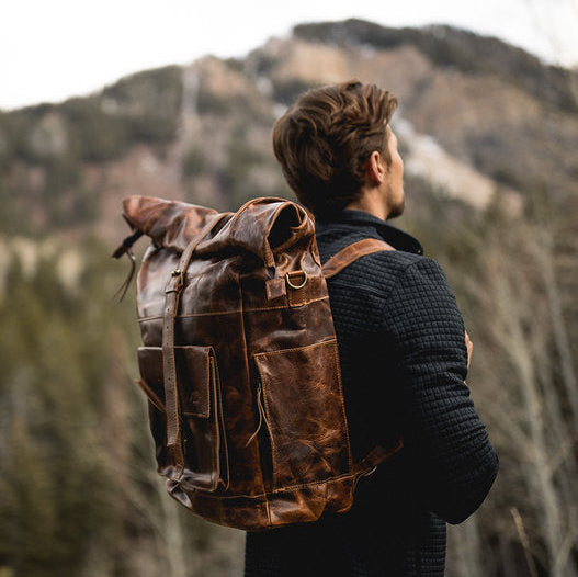 Person with a brown backpack standing in a forest with mountains in the background