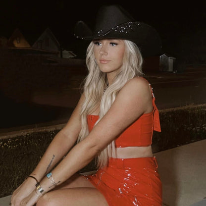 Woman in a red outfit and black cowboy hat sitting on a curb at night.