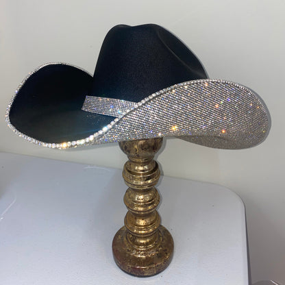 Black cowboy hat with a rhinestone band on a gold stand against a white background